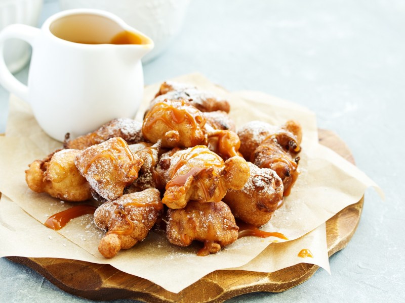 Apfelkrapfen auf einem Holzbrett. Daneben steht eine Kanne mit Glasur.