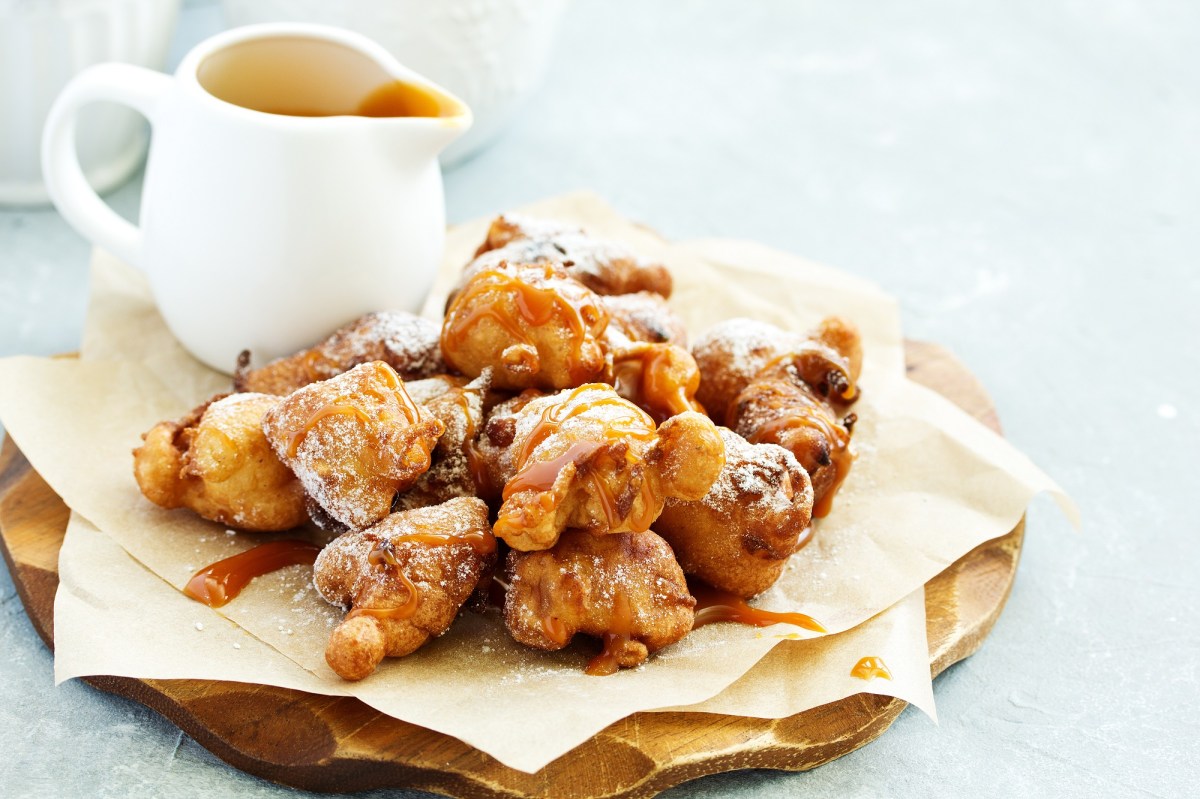 Apfelkrapfen auf einem Holzbrett. Daneben steht eine Kanne mit Glasur.