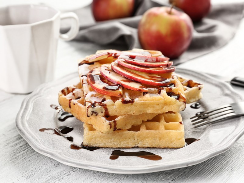 Apfelwaffeln mit SchokosoÃŸe und Apfelscheiben auf einem weiÃŸen Teller. Dahinter liegt ein Apfel.