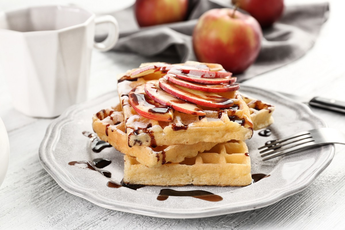 Apfelwaffeln mit SchokosoÃŸe und Apfelscheiben auf einem weiÃŸen Teller. Dahinter liegt ein Apfel.