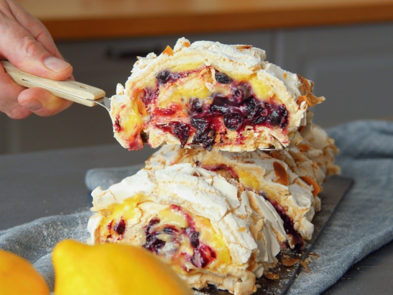 Ein Stück gefüllte Baiserrolle mit Lemon Curd und Pflaumenkompott wird mit einem Tortenheber entnommen.