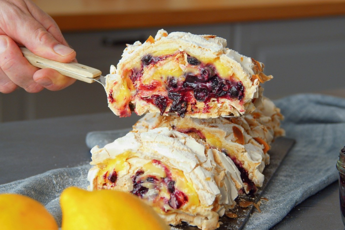 Ein Stück gefüllte Baiserrolle mit Lemon Curd und Pflaumenkompott wird mit einem Tortenheber entnommen.