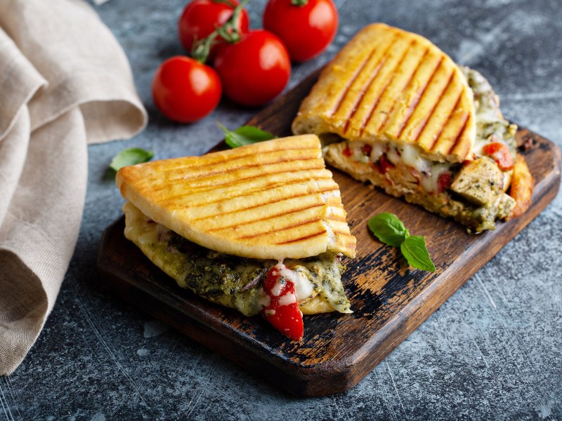 2 Italienische Hähnchen-Paninis mit grünem Pesto und Mozzarella auf einem Brett, im Hintergrund Tomaten.