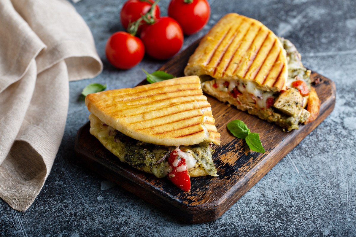 2 Italienische HÃ¤hnchen-Paninis mit grÃ¼nem Pesto und Mozzarella auf einem Brett, im Hintergrund Tomaten.