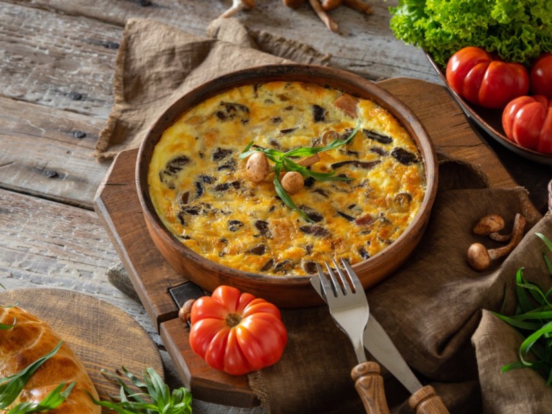 Eine Kartoffel-Frittata mit Pilzen in einer Pfanne. Drumherum liegen frische Zutaten.