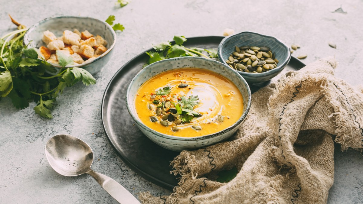 Eine SchÃ¼ssel KÃ¼rbis-Curry-Suppe mit Kernen, CroÃ»tons und KrÃ¤utern verfeinert, daneben je eine SchÃ¼ssel mit Kernen und CroÃ»tons.