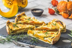 4 Stücke Kürbis-Quiche mit Tofu auf einer Schieferplatte, im Hintergrund verschiedene Kürbisse.