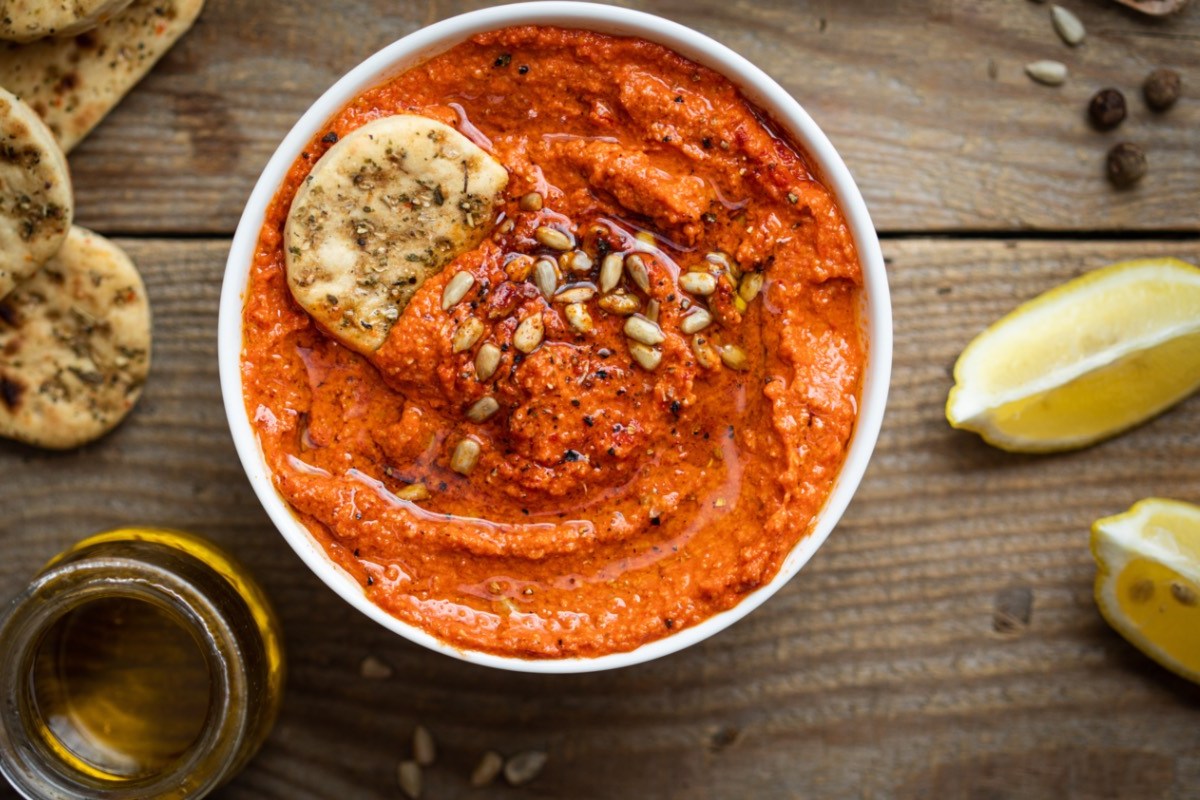 Eine Schale mit Paprika-Tomaten-Hummus. Daneben liegen frische Zutaten.