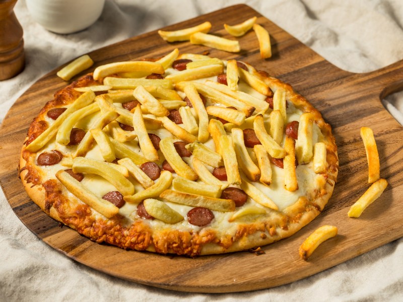 Eine Pizza Americana mit Pommes und Wiener WÃ¼rstchen auf einem Holzbrett.