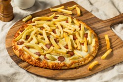 Eine Pizza Americana mit Pommes und Wiener Würstchen auf einem Holzbrett.