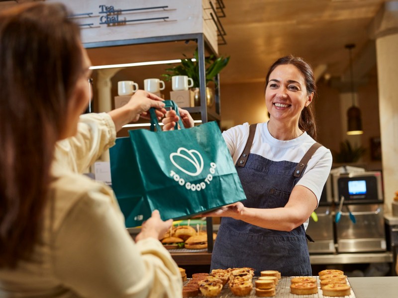 Eine dunkelhaarige Frau reicht einer anderen in einem CafÃ© eine TÃ¼te mit der Aufschrit 