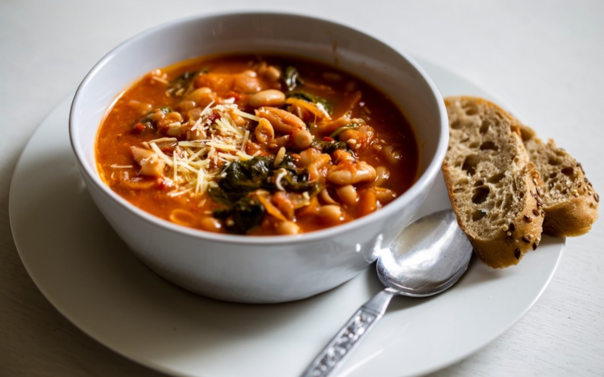 Eine SchÃ¼ssel mit toskanischer Bohnensuppe. Daneben liegen eine Scheibe Brot und LÃ¶ffel.