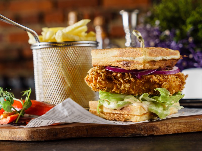 Ein Waffel-Burger belegt mit Hähnchen, Salat, Gewürzgurke und roter Zwiebel, daneben ein Korb mit Pommes.