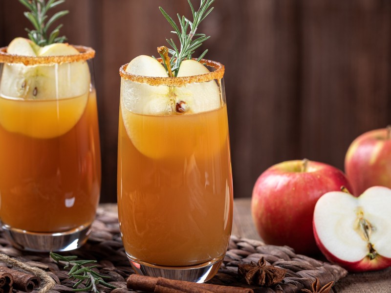 Herbst-Cocktail in zwei GlÃ¤sern, garniert mit Apfel und Rosmarin. Daneben halbierte und ganze Ã„pfel.