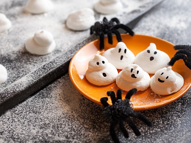 Fünf süße Baiser-Geister auf einem orangen Teller. Spinnen als Halloween-Deko liegen daneben.