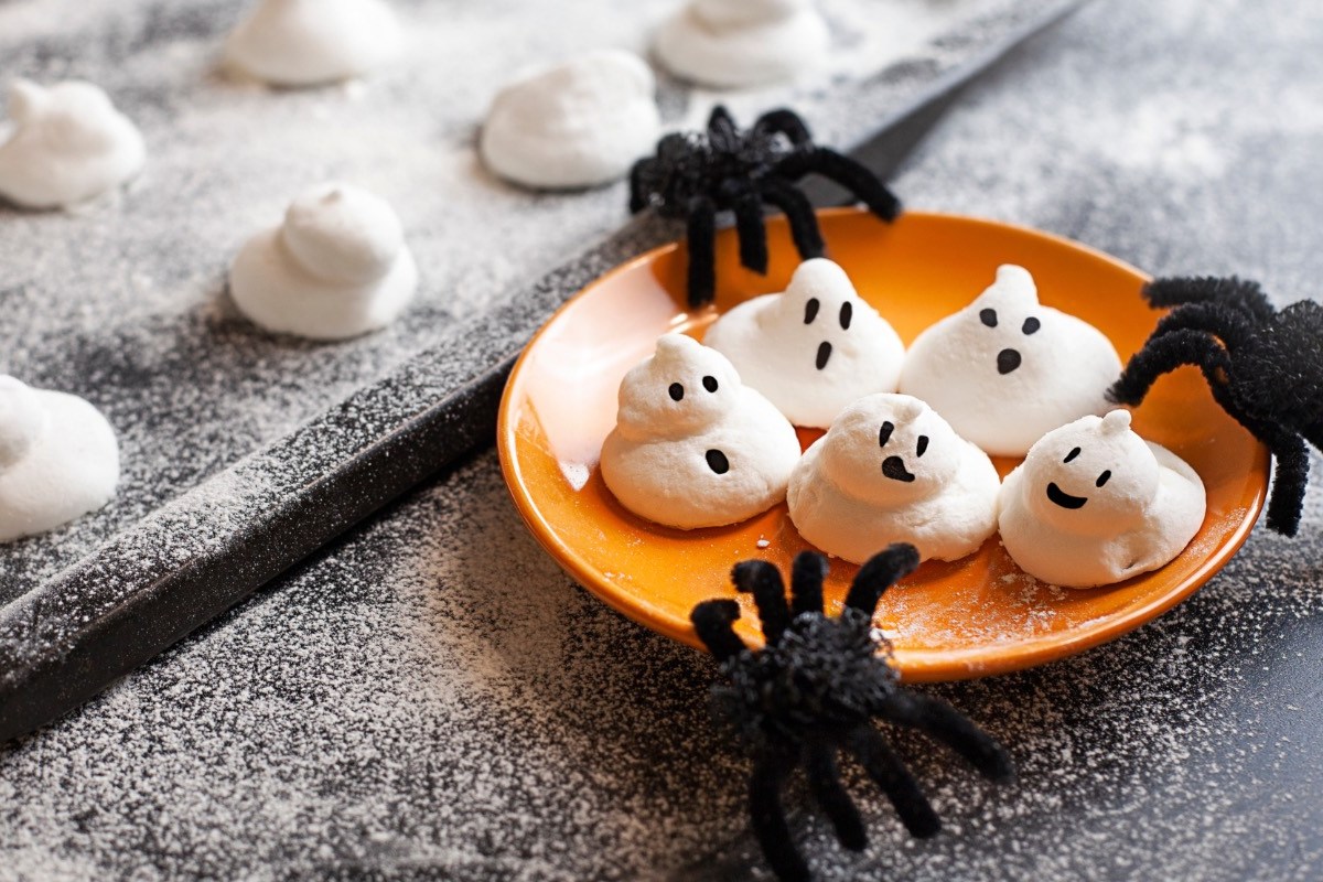 Fünf süße Baiser-Geister auf einem orangen Teller. Spinnen als Halloween-Deko liegen daneben.