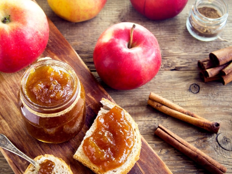 Ein Glas Bratapfelmarmelade. Daneben frische Ã„pfel und ein Brot mit Marmelade bestrichen.