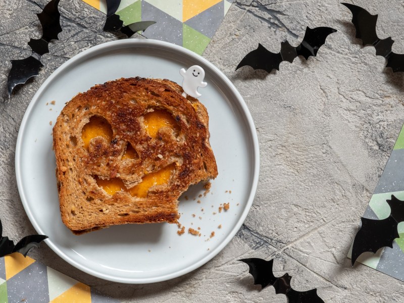 Halloween-Sandwich mit KÃ¤se und KÃ¼rbisgesicht auf einem Teller in der Draufsicht, drumherum Halloween-Deko.