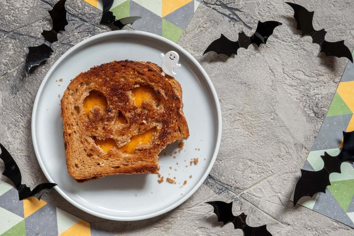 Halloween-Sandwich mit KÃ¤se und KÃ¼rbisgesicht auf einem Teller in der Draufsicht, drumherum Halloween-Deko.