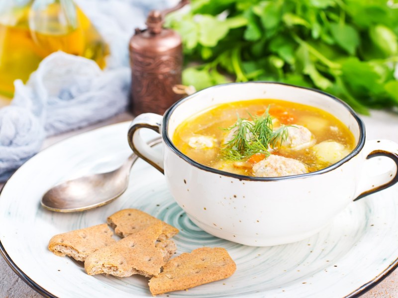 Eine SchÃ¼ssel Hochzeitssuppe mit KlÃ¶ÃŸen und GemÃ¼se auf einem Teller, daneben ein LÃ¶ffel und Cracker, im Hintergrund KrÃ¤uter.