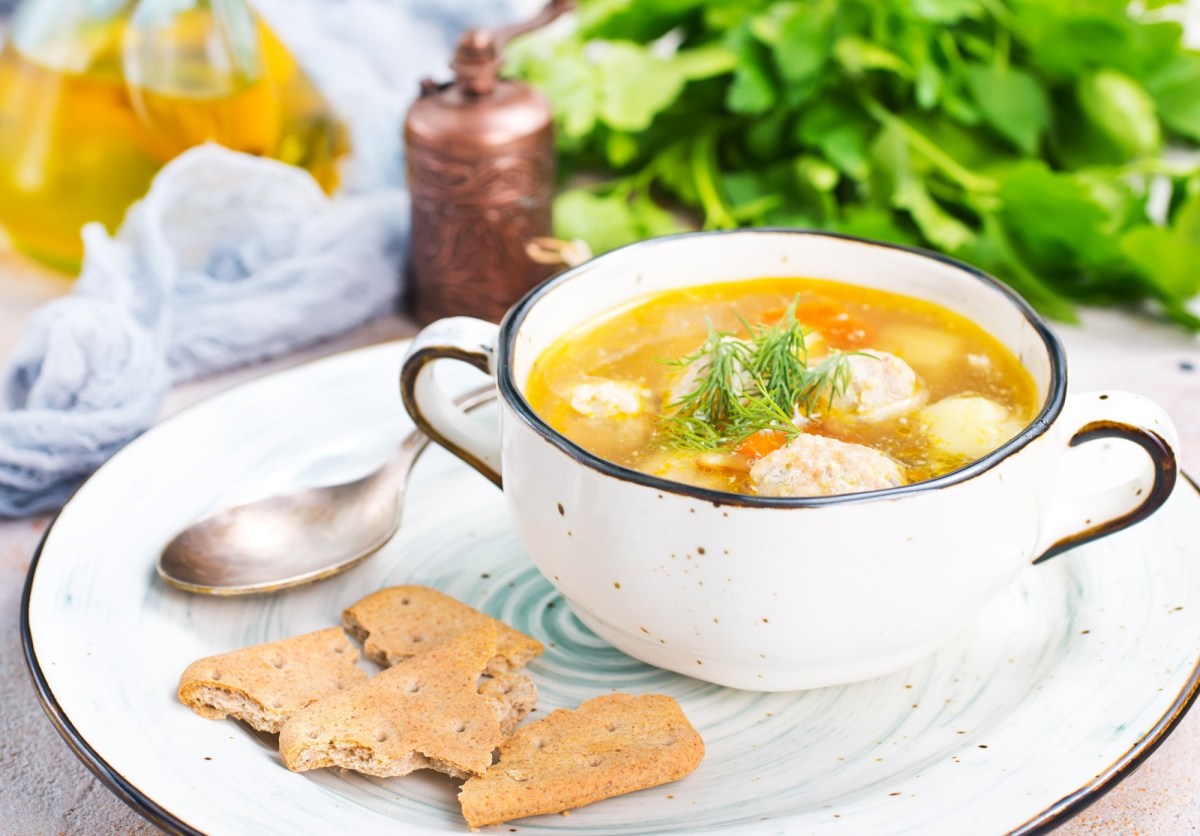 Eine SchÃ¼ssel Hochzeitssuppe mit KlÃ¶ÃŸen und GemÃ¼se auf einem Teller, daneben ein LÃ¶ffel und Cracker, im Hintergrund KrÃ¤uter.