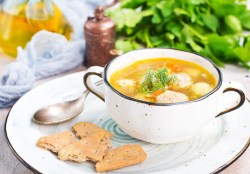 Eine Schüssel Hochzeitssuppe mit Klößen und Gemüse auf einem Teller, daneben ein Löffel und Cracker, im Hintergrund Kräuter.