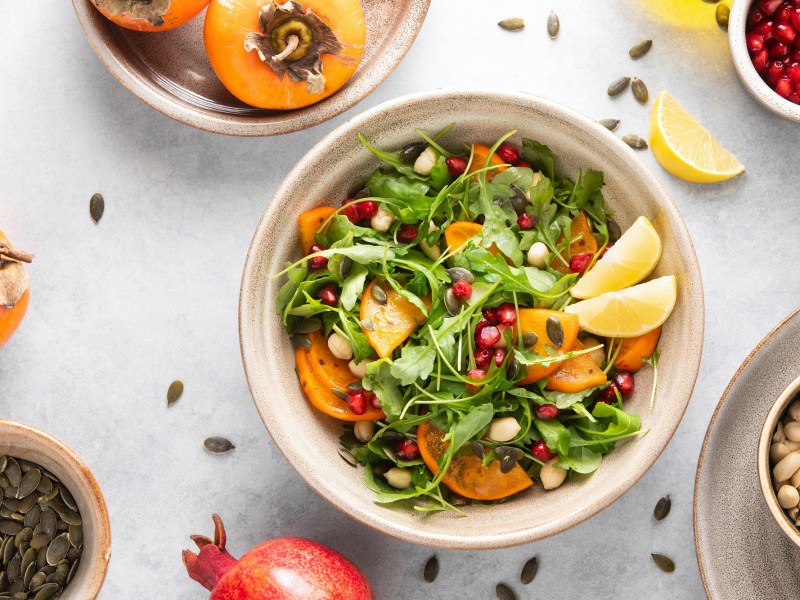 Kaki-Salat mit Rucola in einer weiÃŸen Schale. Daneben liegen ein Granatapfel, NÃ¼sse und Kerne.