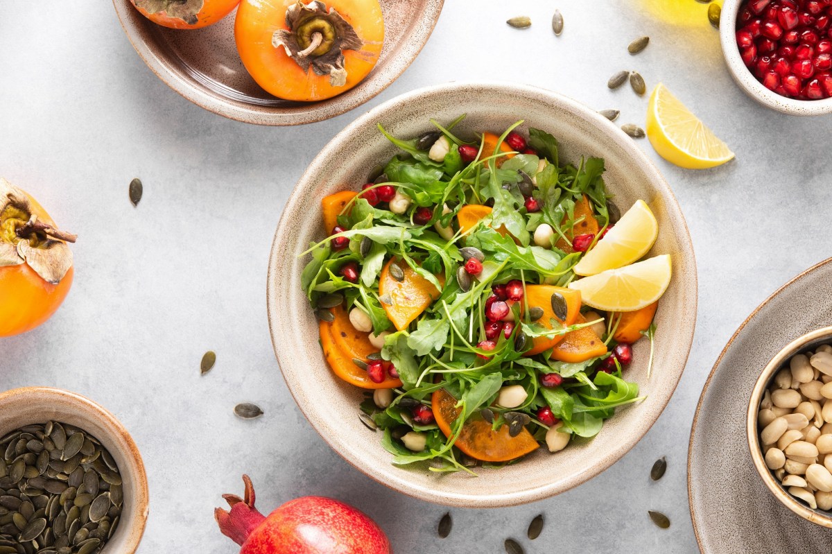 Kaki-Salat mit Rucola in einer weiÃŸen Schale. Daneben liegen ein Granatapfel, NÃ¼sse und Kerne.