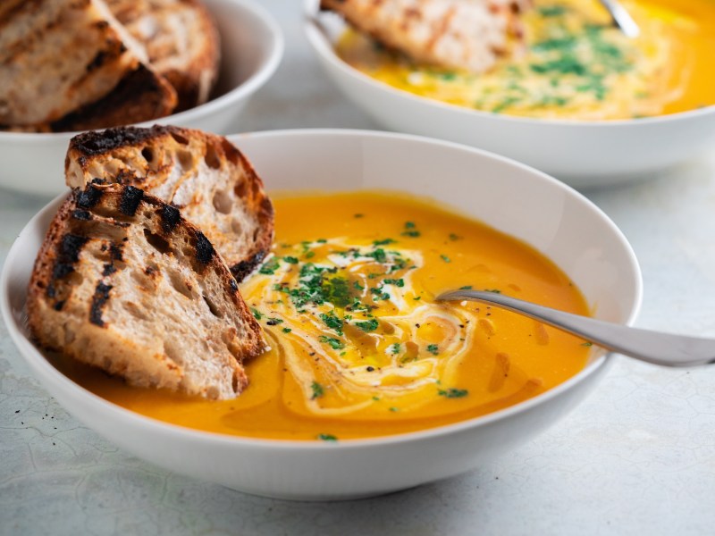 Eine SchÃ¼ssel Kokos-KÃ¼rbissuppe mit Orangensaft und Brotscheiben.