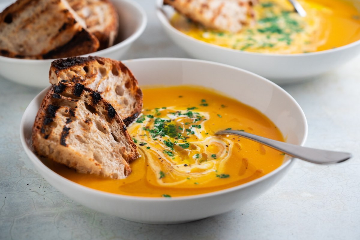 Eine SchÃ¼ssel Kokos-KÃ¼rbissuppe mit Orangensaft und Brotscheiben.