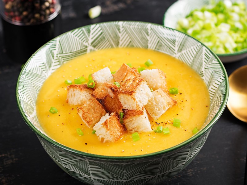 Eine SchÃ¼ssel KÃ¼rbis-Kartoffel-Suppe mit CroÃ»tons und FrÃ¼hlingszwiebel, daneben ein LÃ¶ffel und eine Schale FrÃ¼hlingszwiebel.