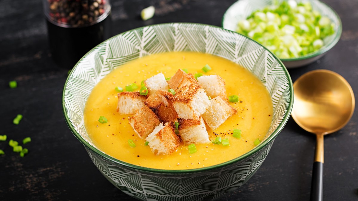 Eine SchÃ¼ssel KÃ¼rbis-Kartoffel-Suppe mit CroÃ»tons und FrÃ¼hlingszwiebel, daneben ein LÃ¶ffel und eine Schale FrÃ¼hlingszwiebel.