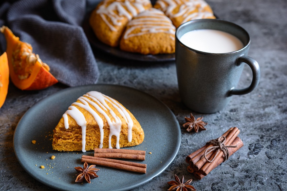 Ein KÃ¼rbis-Scones verziert mit Zuckerguss auf einem Teller, daneben Zimtstangen und eine Tasse Milch, im Hintergrund weitere Scones und KÃ¼rbisspalten.