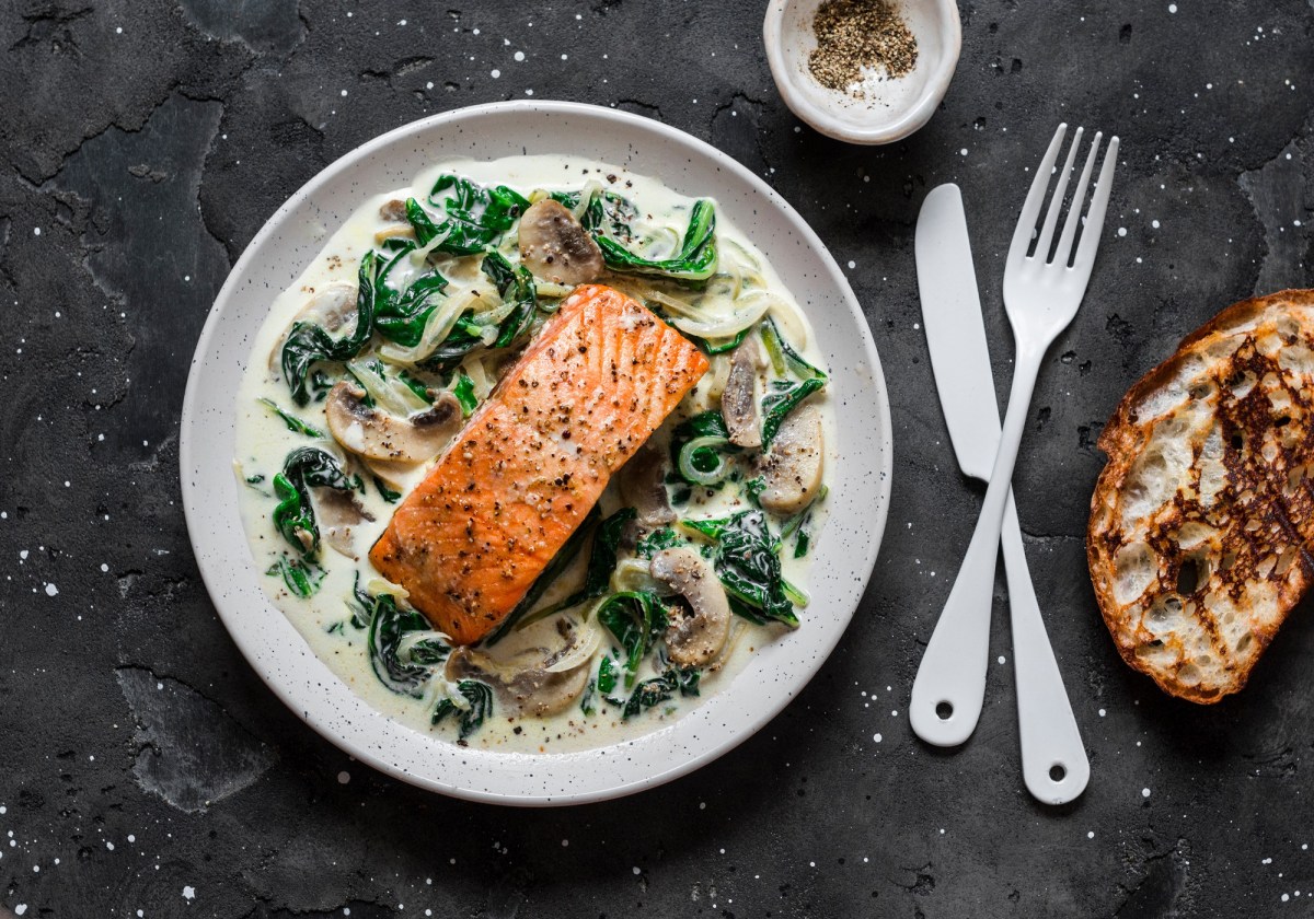 Lachs mit Spinat und Pilzen auf einem weiÃŸen Teller. Daneben liegt Besteck und eine Scheibe gerÃ¶stetes Brot.