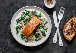 Lachs mit Spinat und Pilzen auf einem weißen Teller. Daneben liegt Besteck und eine Scheibe geröstetes Brot.