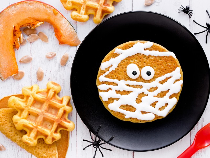 Mumienpfannkuchen mit Zuckerguss auf einem schwarzen Teller, umgeben von Halloween-Dekoration.