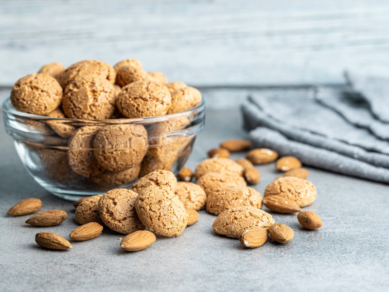 Eine Glasschüssel Amaretti-Kekse, davor einige Kekse und Mandeln.