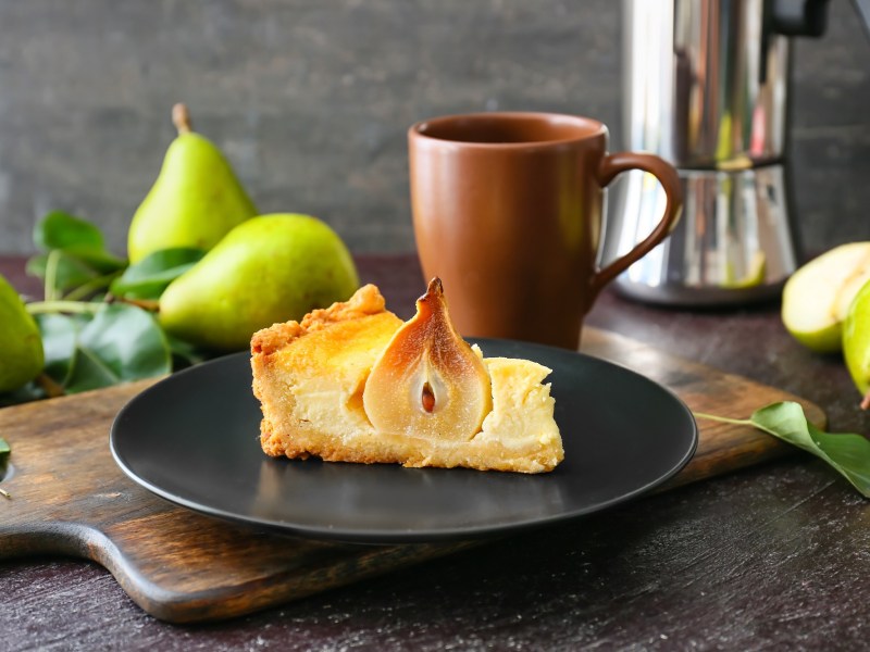 Ein StÃ¼ck Birnen-Schmand-Kuchen mit einer halben Birne auf einem dunklen Teller.