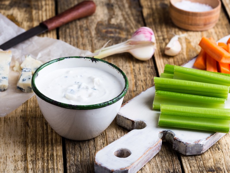 Eine Schale BlauschimmelkÃ¤se-Dressing, daneben GemÃ¼se-Sticks und ein angeschnittener KÃ¶se sowie Knoblauch.