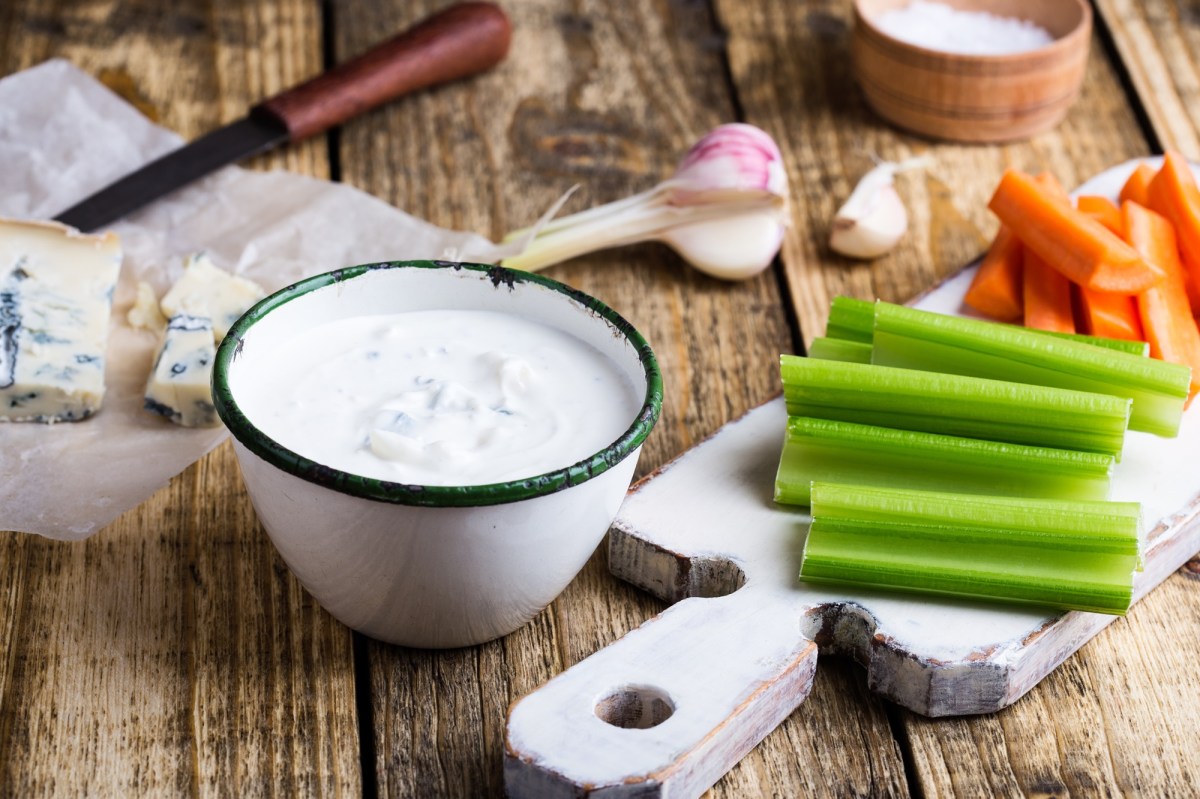 Eine Schale BlauschimmelkÃ¤se-Dressing, daneben GemÃ¼se-Sticks und ein angeschnittener KÃ¶se sowie Knoblauch.