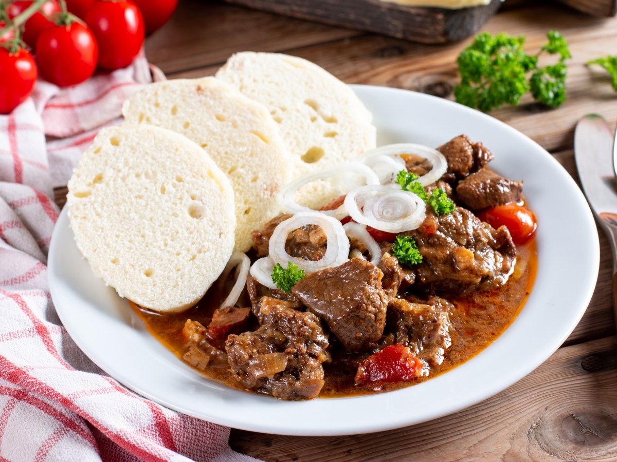 Ein Teller mit Gulasch und böhmischen Knödeln, daneben Besteck und ein kariertes Geschirrtuch.