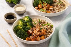 2 Brokkoli-Quinoa-Bowls mit gebratenem Tofu, daneben eine Schale Dressing.