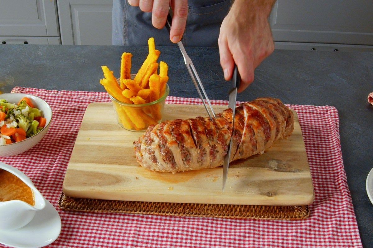 Ein gefÃ¼llter Schweinebraten wird mit einem Messer angeschnitten. Daneben Maispommes.