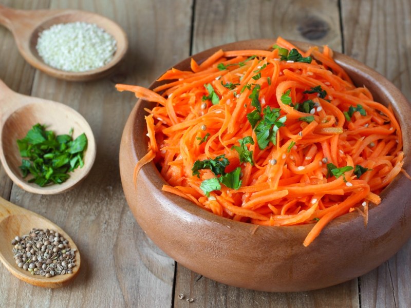 Eine Schüssel koreanischer Karottensalat. Daneben liegen Holzlöffel mit Gewürzen.