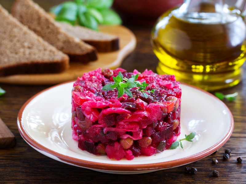 Rote-Bete-Tatar auf einem weiÃŸen Teller, dahinter eine Flasche Ã–l und Brotscheiben.