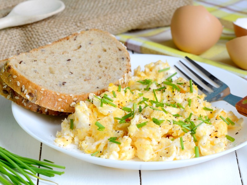 Ein Teller Rührei mit Crème fraîche und zwei Scheiben Brot, daneben Schnittlauch und Eierschalen sowie eine Gabel.