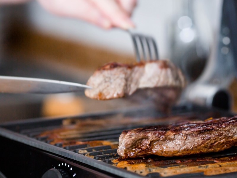 Amazon Black Friday Tefal OptiGrill Kontaktgrill: 2 StÃ¼ck Fleisch auf einem Kontaktgrill, eins wird mit Messer und Gabel weggenommen. Nahaufnahme.