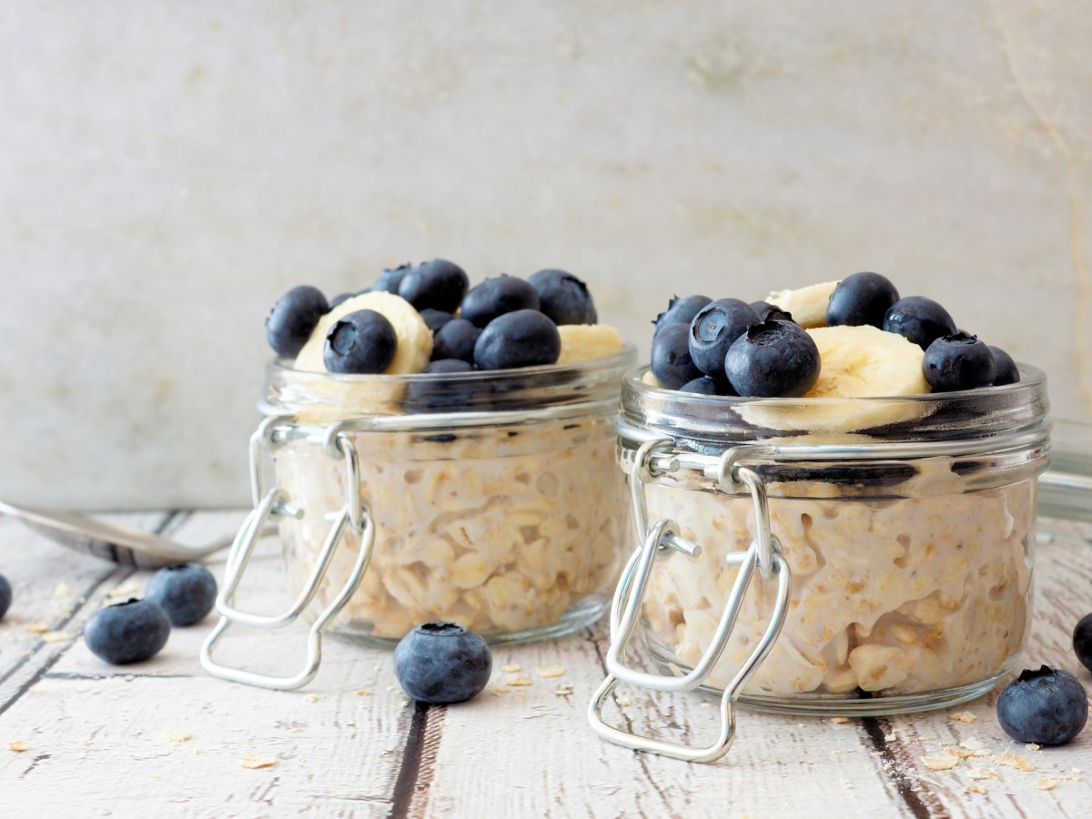 2 GlÃ¤ser Overnight Oats garniert mit Blaubeeren und Banananscheiben, drumherum liegen einige Blaubeeren.