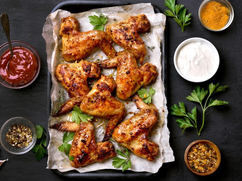 Honig Chicken Wings auf einem mit Backpapier ausgeschlagenen Backblech. Daneben stehen SchÃ¼sseln mit SoÃŸe.