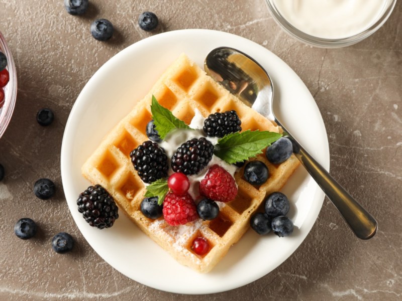 Eine Käsekuchenwaffel mit Früchten, Puderzucker und Sahne auf einem Teller in der Draufsicht.